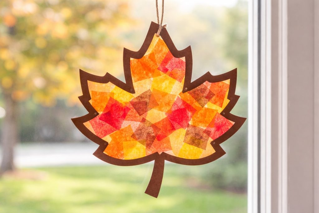 Fall leaf suncatcher made from contact paper and colorful tissue paper hanging on a window, perfect no-mess preschool Thanksgiving craft idea