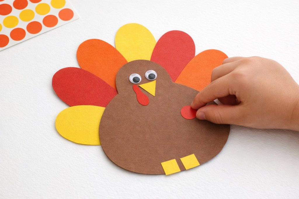 Child's hand placing a red circle sticker on a brown paper turkey shape for an easy preschool Thanksgiving craft idea
